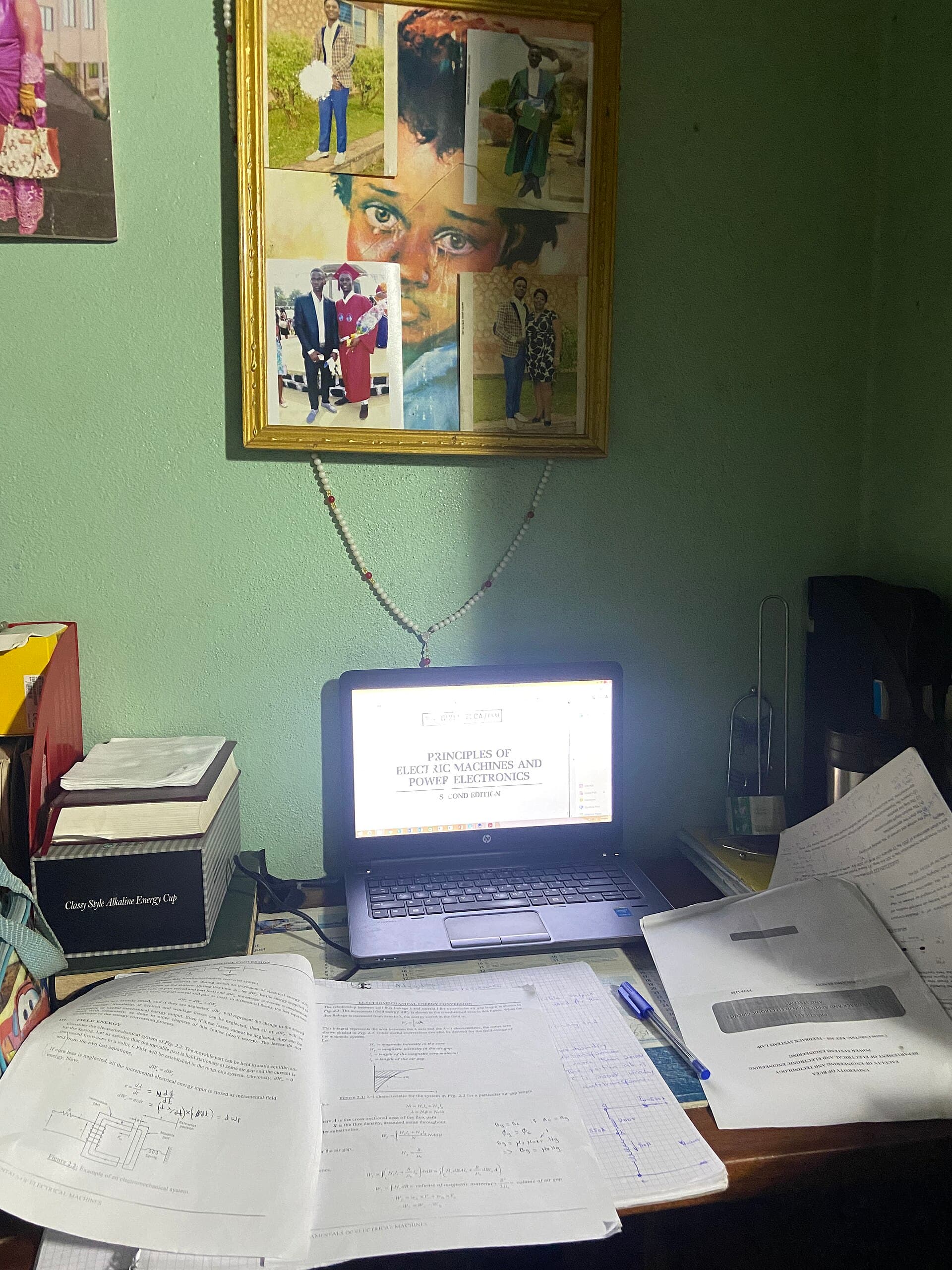 A study table at home with a laptop, printed notes and papers under a framed photo collage
